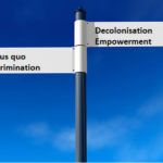 An image of two signs at a crossroads with blue sky in the background. Left is Status que and right is empowerment and decolonisation.